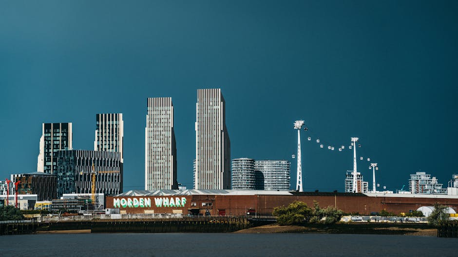 A panoramic view of the Morden Wharf area in London, featuring a cluster of modern high-rise buildings with glass facades and vertical striping, set against a dark, cloudy sky. In the foreground, there is a low-rise brick building with a sign reading 'MORDEN WHARF,' situated near the water's edge, which appears to be part of a site undergoing a home relocation or construction project. To the right, there are several tall floodlights and cranes, indicating ongoing development or industrial activity related to the area. The scene is illuminated by natural daylight, highlighting the structural details of the buildings and surrounding environment, with some greenery present along the water's edge. This setting exemplifies urban development and the logistics involved in house removals or furniture transport within Morden's waterways and commercial zones. Man and Van Morden occasionally operates in this context, supporting packing, loading, and transport services for house removals in the vicinity.