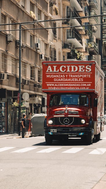 A red moving truck with the branding 'ALCIDES' and web address 'www.alcidesmudanzas.com.ar' is positioned on a city street, partially blocking a pedestrian crossing. The truck is a classic Mercedes-Benz model and is parked close to a multi-storey residential building with balconies featuring plants and outdoor furniture. Nearby, a person dressed in dark clothing stands next to a large grey trash bin, while another individual walks on the sidewalk. The scene includes urban elements such as street lamps, a glass storefront, and air conditioning units attached to the building facades. The loading process appears to involve the transport of household furniture and boxes, typical of house removals or home relocation activities, and the environment suggests an active moving operation within a dense urban area, consistent with professional removals services as offered by companies like Man and Van Morden.