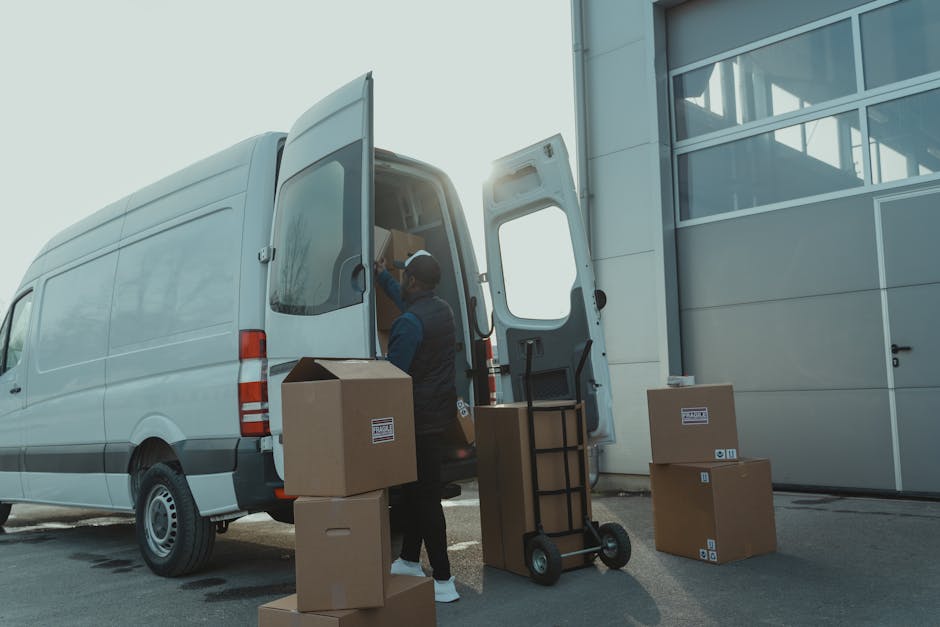 A man wearing a dark jacket and a cap is loading cardboard boxes into the open back of a large white van parked outside a modern building with large glass windows. Several packed boxes are positioned on the ground and a collapsible hand truck is nearby, ready for use. The boxes are sealed with packing tape and feature labels, indicating organized packing for a home relocation or moving process. The scene is well-lit with natural daylight, and the setting appears to be a driveway or designated loading area adjacent to the building. This image captures the logistics of furniture transport and packing as part of a professional removal service, representing the loading process typical in house removals managed by [COMPANY_NAME], such as Man and Van Morden, for clients in the Morden SM4 area.