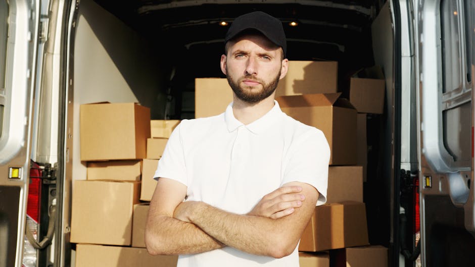 A man wearing a black cap and a white polo shirt stands with his arms crossed inside the open rear of a van loaded with numerous cardboard boxes of various sizes, some wrapped in plastic. The boxes are stacked neatly in the vehicle, which is parked on a paved driveway. The interior of the van is partially visible, showing a spacious cargo area prepared for a house removal. The image captures the process of furniture transport and packing during a home relocation, illustrating the careful loading process handled by professional removals services such as Man and Van Morden.