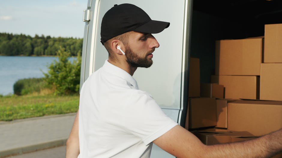 A man wearing a dark jacket and a cap is loading cardboard boxes into the open back of a large white van parked outside a modern building with large glass windows. Several packed boxes are positioned on the ground and a collapsible hand truck is nearby, ready for use. The boxes are sealed with packing tape and feature labels, indicating organized packing for a home relocation or moving process. The scene is well-lit with natural daylight, and the setting appears to be a driveway or designated loading area adjacent to the building. This image captures the logistics of furniture transport and packing as part of a professional removal service, representing the loading process typical in house removals managed by [COMPANY_NAME], such as Man and Van Morden, for clients in the Morden SM4 area.