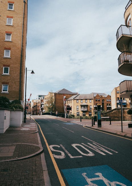 A street view capturing a narrow city road with a sloped incline, surrounded by multi-storey residential buildings made of brick and concrete. The road features a 'SLOW' road marking in white paint, w