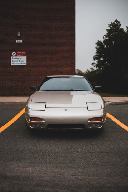 A silver sports car, positioned head-on in a parking space marked with yellow lines, is parked directly facing a brick building with a small sign indicating parking restrictions. The car features a sleek, low-profile design typical of home relocation or furniture transport vehicles, with covered headlights and a streamlined shape. Behind the car, there is a concrete sidewalk bordering the parking area, and to the right, a few tall trees with dark green foliage are visible under a cloudy sky. The scene is captured during daytime with diffuse lighting, emphasizing the car’s smooth metallic surface. This setup suggests readiness for a furniture transport or moving service, as provided by companies like Man and Van Morden, for house removals or home relocation processes in Morden. The parking lot environment and the parked vehicle imply a preparatory stage for loading or unloading household belongings in a secure, designated space.
