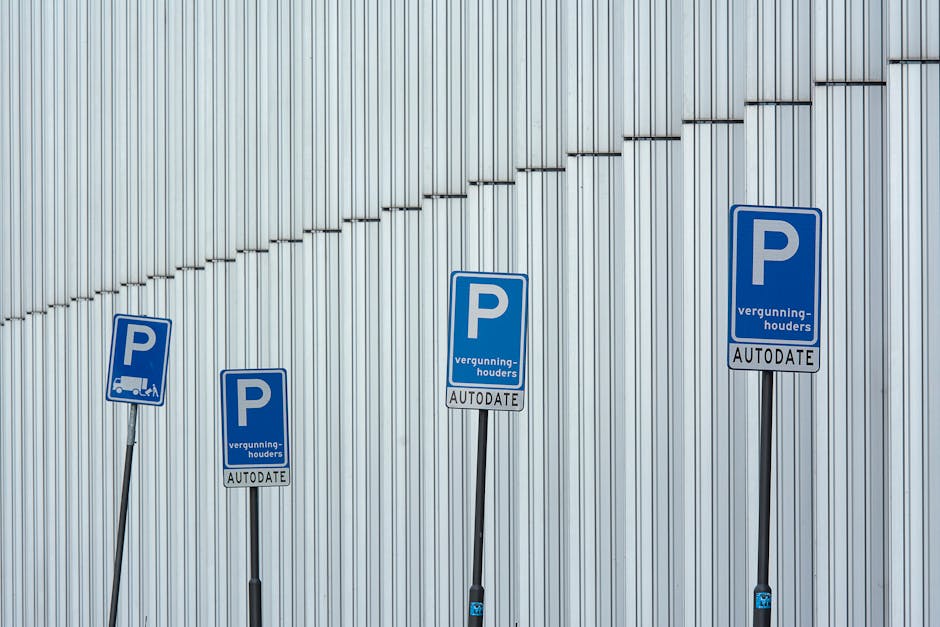 Four blue parking permit signs mounted on black posts are positioned in front of a modern, silver metallic wall with vertical ridges. The signs feature a white 'P' symbol and additional text, indicating parking restrictions with the word 'vergunninghouders' and 'AUTODATE' at the bottom. The signs are placed at varying heights along a paved area or sidewalk, with no vehicles or moving objects visible. The environment appears to be an outdoor urban or industrial setting, possibly near a commercial building or parking lot entrance, with soft lighting illuminating the scene. The image relates to parking permits or regulations, subtly supporting content about house removals or moving logistics within the context of applying for or managing parking permits for moving services, such as those provided by Man and Van Morden.