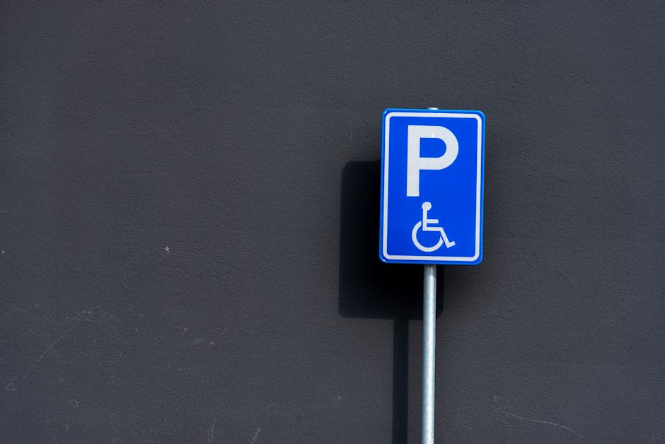 A silver sports car, positioned head-on in a parking space marked with yellow lines, is parked directly facing a brick building with a small sign indicating parking restrictions. The car features a sleek, low-profile design typical of home relocation or furniture transport vehicles, with covered headlights and a streamlined shape. Behind the car, there is a concrete sidewalk bordering the parking area, and to the right, a few tall trees with dark green foliage are visible under a cloudy sky. The scene is captured during daytime with diffuse lighting, emphasizing the car’s smooth metallic surface. This setup suggests readiness for a furniture transport or moving service, as provided by companies like Man and Van Morden, for house removals or home relocation processes in Morden. The parking lot environment and the parked vehicle imply a preparatory stage for loading or unloading household belongings in a secure, designated space.