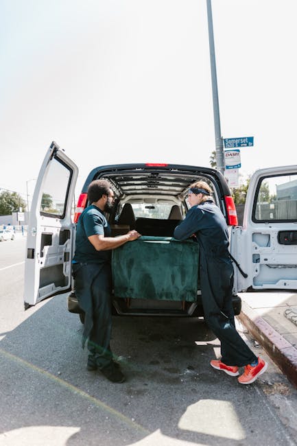 Two movers from Man and Van Morden are loading a large, dark green upholstered piece of furniture into the rear of a black van equipped for furniture transport as part of a home relocation process. The van's rear doors are open, revealing the spacious interior suitable for moving household items. The workers are standing on an outdoor pavement area near a curb, handling the furniture carefully while wearing casual work attire, including gloves. The scene is set during daylight hours with clear weather, and a street sign and other vehicles are visible in the background. This image illustrates professional packing and loading activities typical of house removals services offered in Morden and the surrounding areas.