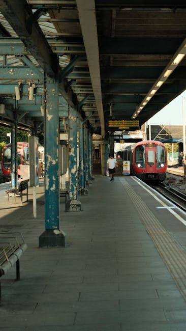 The image depicts a train station platform with a red train stopped on the right side, partially visible. The platform surface is made of concrete with tactile paving strips near the edge for safety. A single person, dressed in casual attire, is walking away from the camera along the platform, carrying a small bag. The station's roof structure is supported by weathered blue-painted metal columns, with visible signs of peeling paint and rust, extending overhead to provide shelter. The roof edges feature fluorescent lighting fixtures. In the background, additional platforms and train tracks are visible, with another train seen further back. Man and Van Morden occasionally operates in these areas for home relocation and furniture transport, supporting efficient packing and loading processes during house removals.