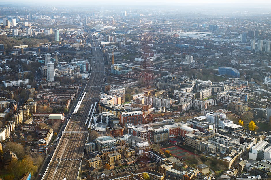 Aerial view of a densely populated urban area with a network of railway tracks running diagonally across the scene. Various residential and commercial buildings of different sizes and architectural styles are visible, including apartment complexes, office buildings, and houses, some with flat roofs and others with pitched roofs. The streets and pathways weave through the cityscape, with trees scattered throughout, some displaying autumn foliage. In the foreground, a section of a property with visible entryway and pavement can be seen, possibly part of a home undergoing relocation or furniture transport. The lighting suggests daytime with clear weather. This image represents a typical city environment where local removals and home moving services, such as those provided by Man and Van Morden, could operate to facilitate house removals, packing, and furniture loading within this urban context.