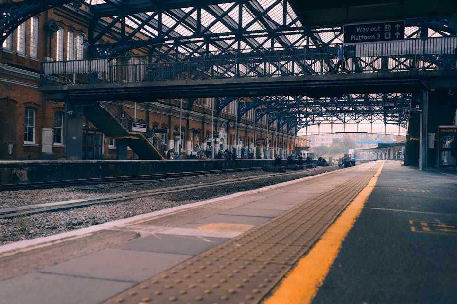 The image depicts a train station platform with a red train stopped on the right side, partially visible. The platform surface is made of concrete with tactile paving strips near the edge for safety. A single person, dressed in casual attire, is walking away from the camera along the platform, carrying a small bag. The station's roof structure is supported by weathered blue-painted metal columns, with visible signs of peeling paint and rust, extending overhead to provide shelter. The roof edges feature fluorescent lighting fixtures. In the background, additional platforms and train tracks are visible, with another train seen further back. Man and Van Morden occasionally operates in these areas for home relocation and furniture transport, supporting efficient packing and loading processes during house removals.