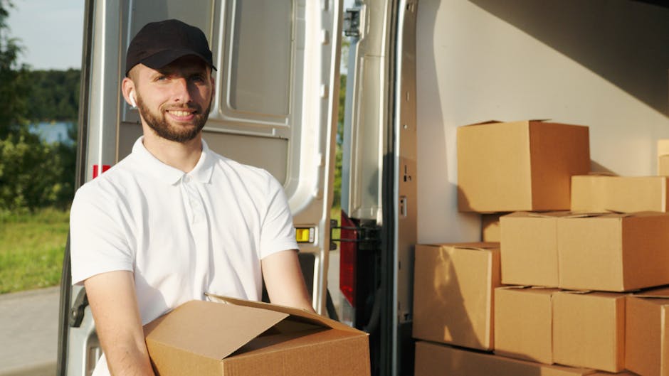 A man wearing a white polo shirt and a black cap is standing outdoors beside an open moving van, holding a medium-sized cardboard box. Inside the van, there are several other cardboard boxes stacked neatly, some covered with packing materials like bubble wrap or plastic wrap. The van's interior is partially visible, showing the loading area with more boxes and possible packing equipment. The background features a green landscape with trees and a blue sky, indicating it's daytime with natural lighting. The scene depicts the process of home relocation, with the man likely involved in furniture transport and packing and moving activities associated with a professional removals service. The environment appears tidy and organized, reflecting a typical house removal operation supported by [COMPANY_NAME], specializing in same-day moving services in Morden.