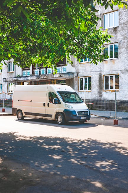 A white moving van parked on a residential street with a concrete multi-storey building in the background. The van is positioned parallel to the pavement, with its side door closed and no visible branding. The area around the van is shaded by a large leafy green tree, casting dappled sunlight on the asphalt. In the foreground, a few wooden bollards line the edge of the parking area, separating it from the footpath. The building behind features multiple windows, some of which display decorative frosted glass patterns, indicating residential flats. The image captures a quiet moment during a home relocation or furniture transport process, typical of professional removals services such as those offered by Man and Van Morden. The scene emphasizes the logistical aspects of loading or unloading furniture and boxes, although no activities are currently visible. The environment appears clean and organized, with natural lighting highlighting the details of the vehicle and surroundings.