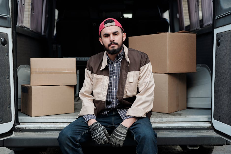 A man wearing a white polo shirt and a black cap is standing outdoors beside an open moving van, holding a medium-sized cardboard box. Inside the van, there are several other cardboard boxes stacked neatly, some covered with packing materials like bubble wrap or plastic wrap. The van's interior is partially visible, showing the loading area with more boxes and possible packing equipment. The background features a green landscape with trees and a blue sky, indicating it's daytime with natural lighting. The scene depicts the process of home relocation, with the man likely involved in furniture transport and packing and moving activities associated with a professional removals service. The environment appears tidy and organized, reflecting a typical house removal operation supported by [COMPANY_NAME], specializing in same-day moving services in Morden.