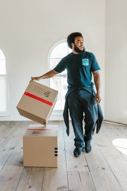 A man with a dark afro hairstyle and beard, dressed in a navy blue T-shirt with a logo and dark trousers, is inside a well-lit room with light-colored walls and a wooden floor. He is holding a cardboard box with red tape and labels, about to place or move it onto another closed cardboard box on the floor. The room features a large, arched window that lets in natural daylight, illuminating the space and highlighting the packaging materials. This scene depicts the packing and loading process involved in a home relocation, with the man's relaxed posture indicating careful handling of the boxes in a typical moving scenario. As part of the furniture transport and packing process, [COMPANY_NAME], such as Man and Van Morden, might be involved in similar tasks, including the careful loading of boxes and furniture into vans for transport. The focus is on the careful management of moving logistics, with an emphasis on safe handling of packed items during a house removal.