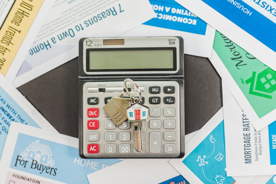 A calculator with a keychain attached, featuring a small house-shaped and a colorful house-shaped charm, rests on top of scattered property-related documents including mortgage forms and home-related brochures. The calculator is a 12-digit electronic device with white and gray buttons, a black frame, and a large rectangular digital display at the top. The documents beneath are in various colors such as blue, green, and yellow, with text related to home buying, mortgage rates, and property transactions. The scene appears to be indoors, on a dark surface, highlighting the context of home ownership and financial planning in relation to house removals or relocation services, as provided by Man and Van Morden, within the scope of a flat move guide for Cannon Hill estate residents.