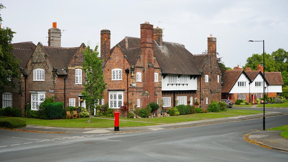 A row of traditional brick residential buildings with pitched roofs and multiple chimneys along a quiet suburban street. The buildings feature white-framed windows, some with decorative patterns, and small garden areas with bushes and trees in front. One house has a white bay window with a prominent balcony above, while another displays a wooden porch. The street includes a red postbox on the pavement, a black streetlamp, and a parked vehicle under a carport. The overall scene captures a peaceful neighborhood typical of Morden, with ambient natural lighting indicating daytime, and minimal traffic. The setting reflects the typical environment for home relocations and house removals, which companies like Man and Van Morden might operate within, while showcasing the local residential architecture and streetscape.