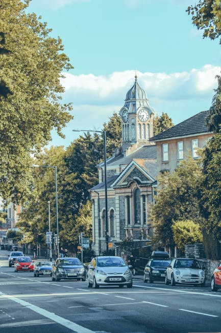 A busy street scene in Morden during daytime with several cars parked along the pavement and in motion, including a white van used for home relocation purposes. In the background, there is a historic clock tower with a pointed spire and ornate architectural details, surrounded by lush green trees and nearby buildings. The image captures the loading process of furniture and boxes onto the van, which is part of a professional removals service provided by Man and Van Morden. Cardboard boxes, some wrapped in plastic for protection, are visible near the entrance of a property, with a trolley and packing materials present as part of the packing and moving operations. The natural daylight and clear sky highlight the urban environment, emphasizing the detailed logistics involved in house removals within Morden, with careful attention to local regulations such as those outlined by Merton Council.